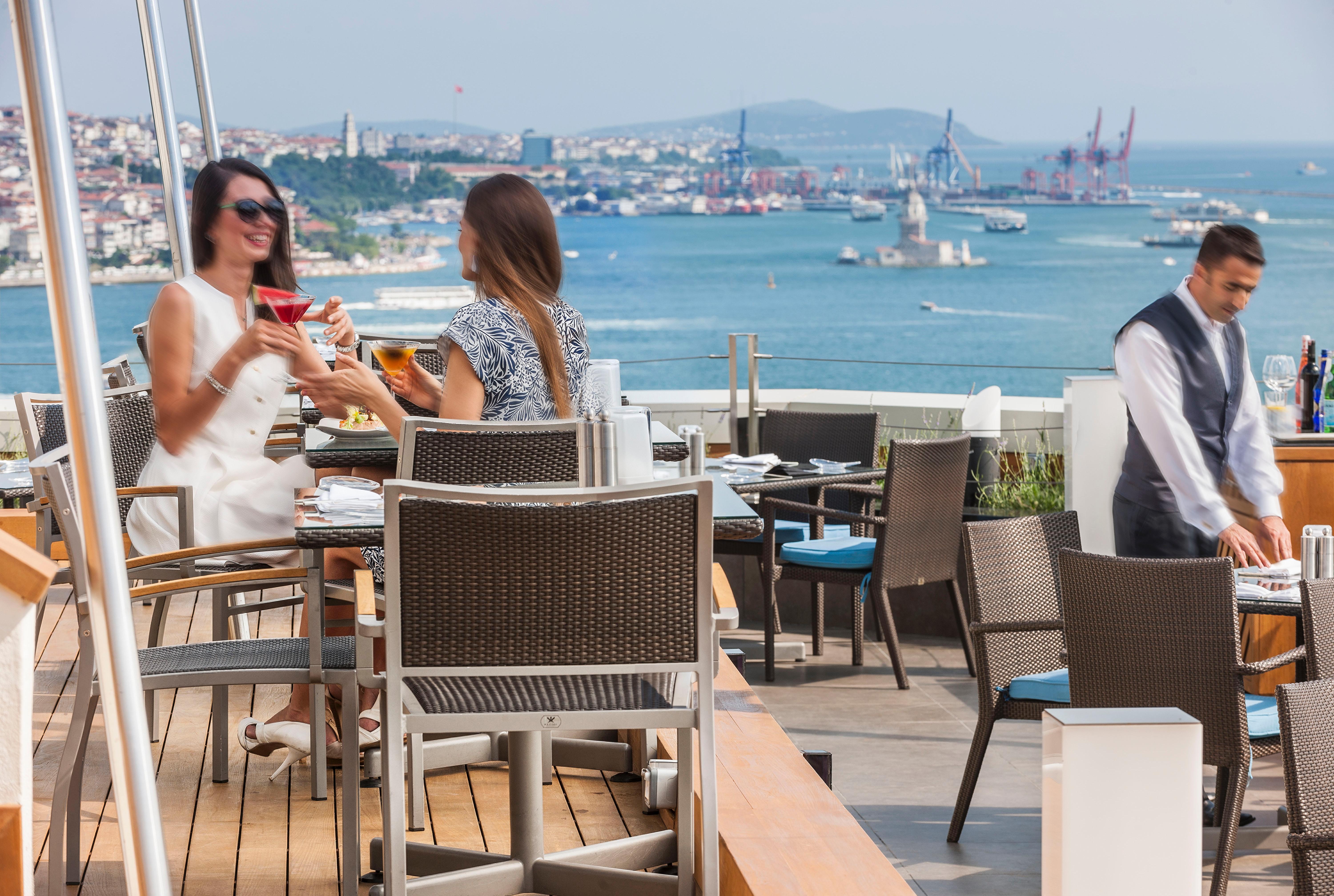 Swissotel The Bosphorus Istanbul Exterior photo The photo depicts a vibrant outdoor setting at a restaurant with a view of the sea and distant cityscape. Two women are seated at a table, enjoying drinks and engaged in conversation, while a waiter in a uniform is nearby. The environment appears rel