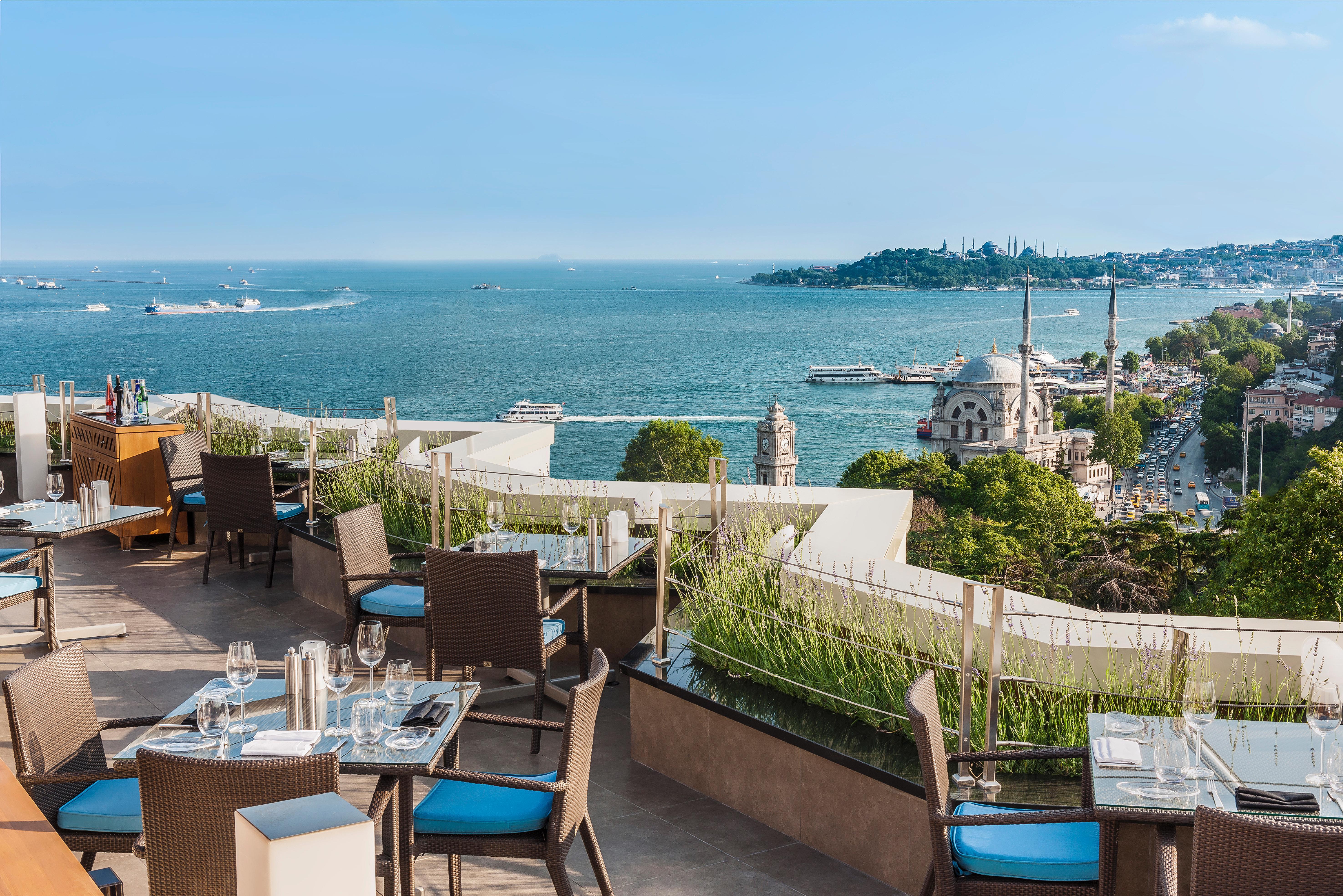 Swissotel The Bosphorus Istanbul Exterior photo The image shows a beautiful outdoor dining area on a terrace overlooking a scenic coastal view. The setup includes several tables with elegant dining arrangements, featuring chairs with blue cushions. In the background, there is a vast body of water,