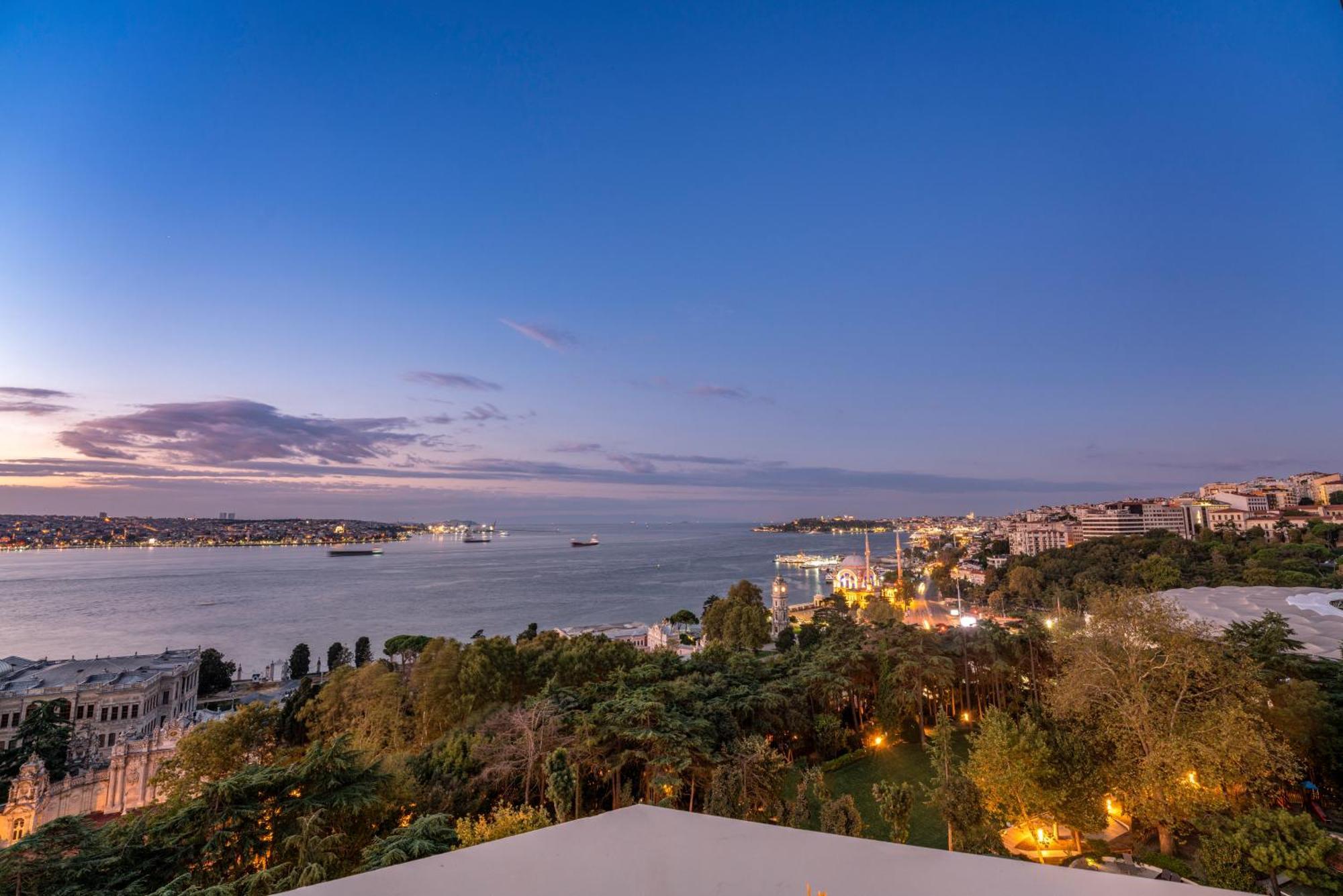 Swissotel The Bosphorus Istanbul Exterior photo The photo shows a scenic view of a waterfront area during twilight. The sky transitions from daylight to dusk, displaying soft hues of blue and hints of purple. In the foreground, there are lush trees and greenery, while the background features a wel