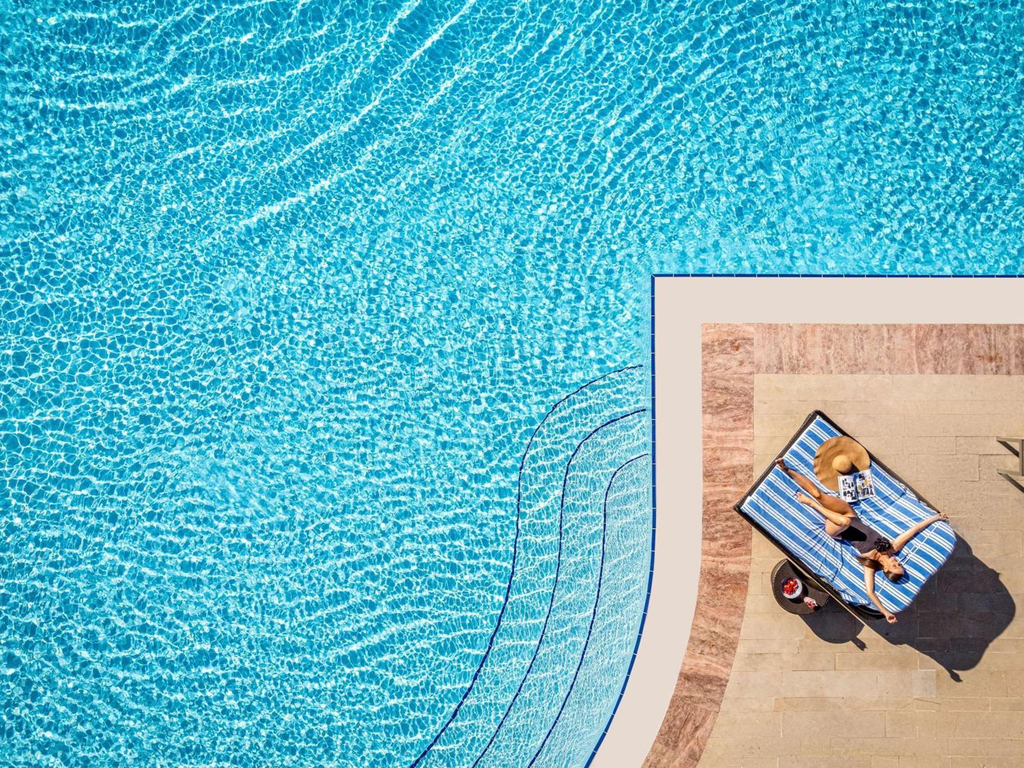 Swissotel The Bosphorus Istanbul Exterior photo The photo shows a clear blue swimming pool with rippling water. At the edge of the pool, there is a lounge chair with a person relaxing on it, surrounded by a wooden deck. The person appears to be enjoying a sunny day, possibly sunbathing. The scene 