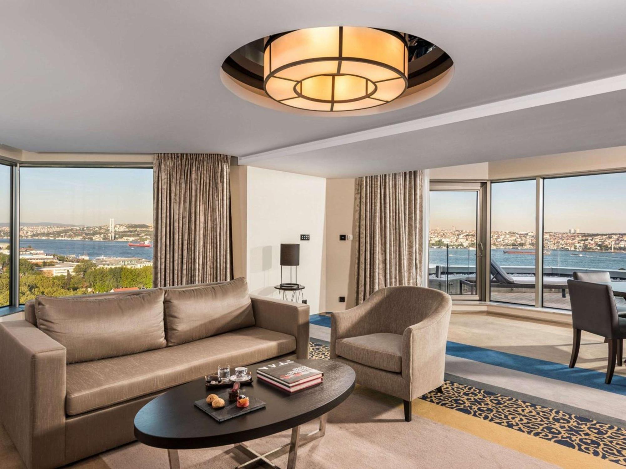 Swissotel The Bosphorus Istanbul Exterior photo The photo shows a stylish hotel room or living space. It features a modern design with a large window offering a view of a body of water in the background. The room includes a comfortable gray sofa and a round chair, arranged around a circular coffee